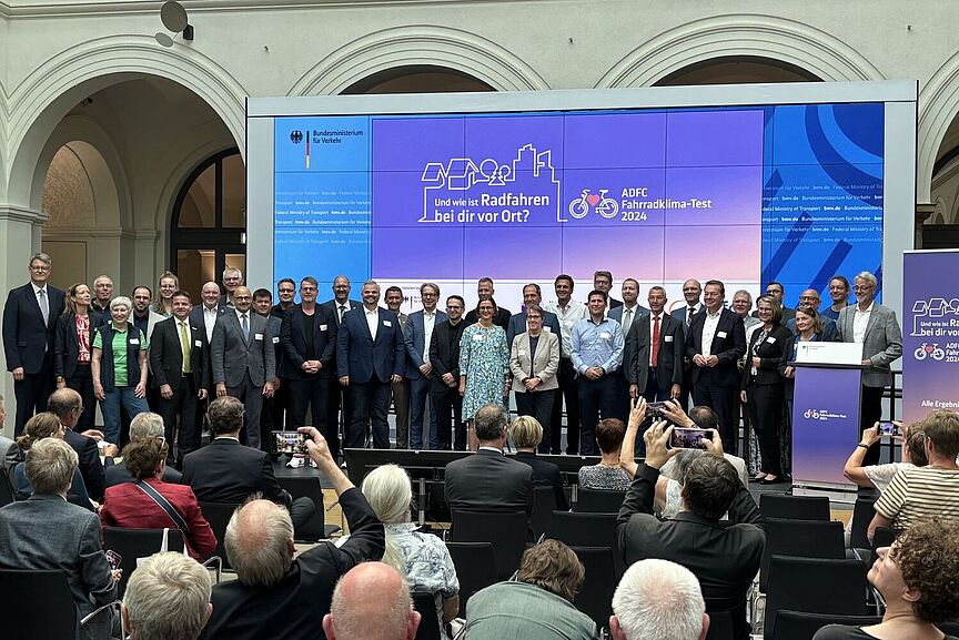 FKT-Präsentation Auf einer großen Bühne im Bundesverkehrsministerium sind zahlreiche Städtevertreterinnen und -vertreter versammelt, eingerahmt von Bundesverkehrsminister Schnieder und ADFC-Bundesvorsitzenden Masurat.